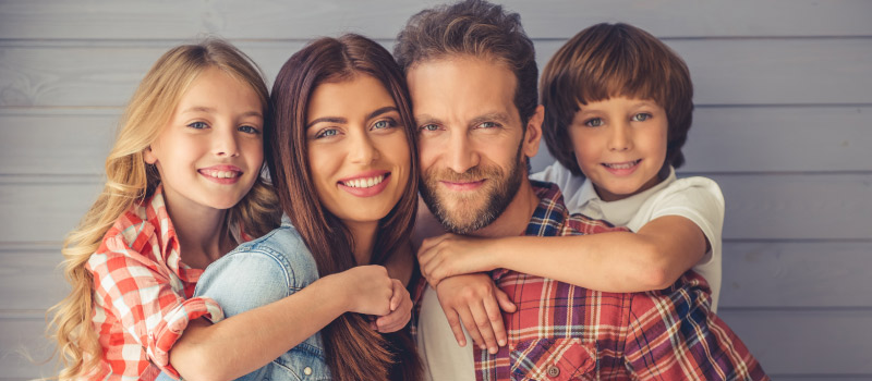 A daughter and son embrace their mom and dad from behind as they smile against a gray wall after receiving family dental care in Liberty, MO