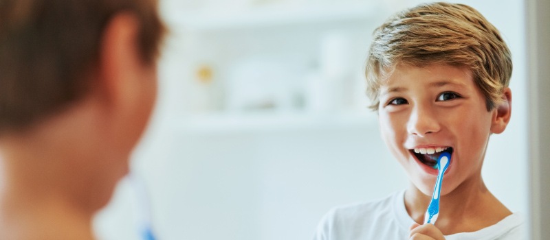 Blonde boy smiles at himself in the mirror as he brushes his teeth with a soft-bristled toothbrush