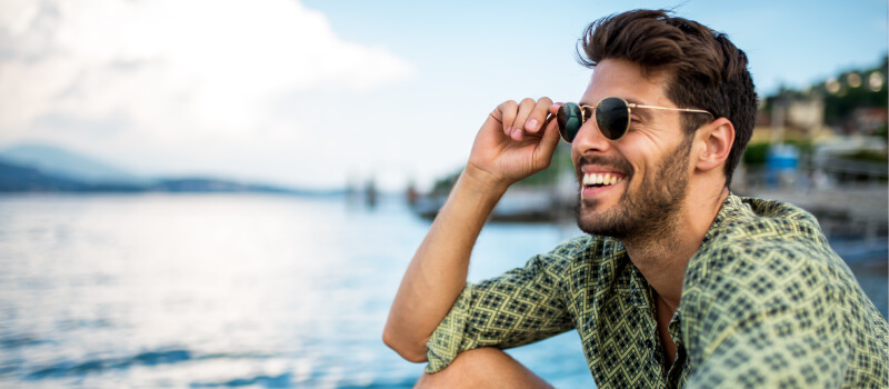 Brunette man wearing sunglasses smiles at the lake after restoring his smile with dental implants