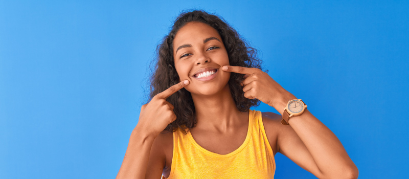 Brunette woman against a blue wall in a yellow tanktop points to her smile with a dental crown in Liberty, MO