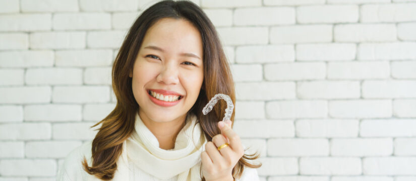 Brunette woman in a big sweater smiles while holding her clear aligners