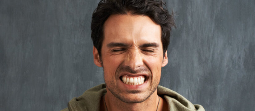 Brunette man clenches his teeth against a gray background while wearing a green hoodie