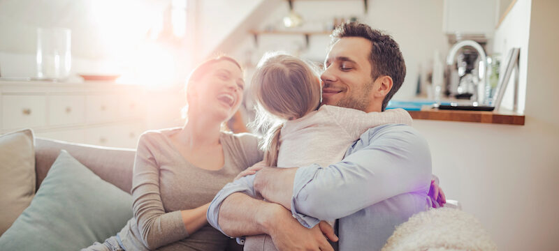 Closeup of a mom and dad smiling as they embrace their daughter on the couch in their living room