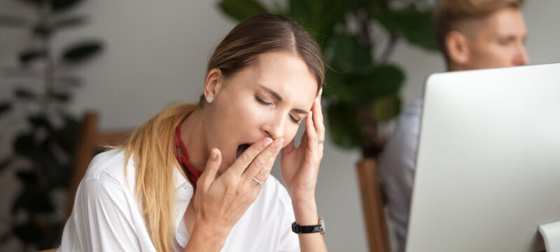 Closeup of a woman in a white blouse yawning at her computer because she has daytime sleepiness and fatigue from sleep apnea