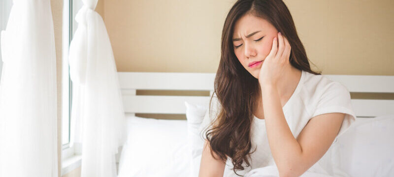 Asian woman in a white shirt cringes in pain and touches her cheek due to sensitive teeth