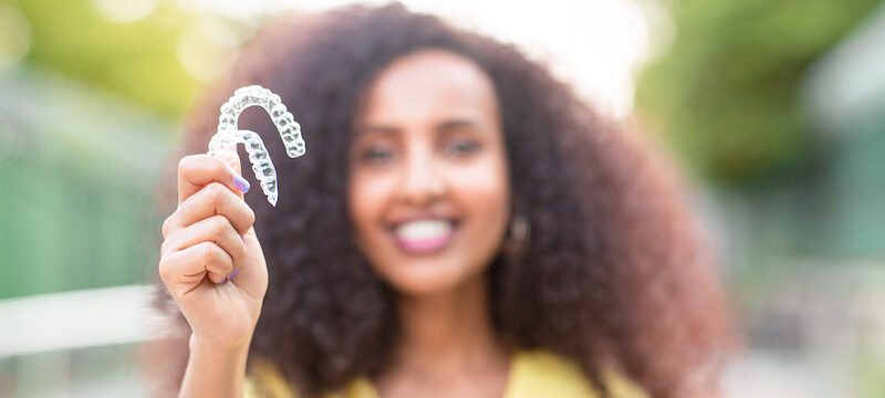 Blurred Black woman in a yellow blouse smiles as she holds up two Invisalign clear aligners outside